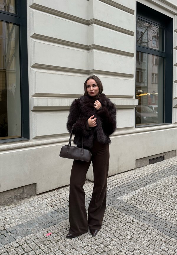 Femme debout sur une rue pavée, portant une veste en fourrure foncée, un pantalon à jambes larges, des chaussures pointues, et tenant un petit sac à main en cuir près d’une façade de bâtiment.