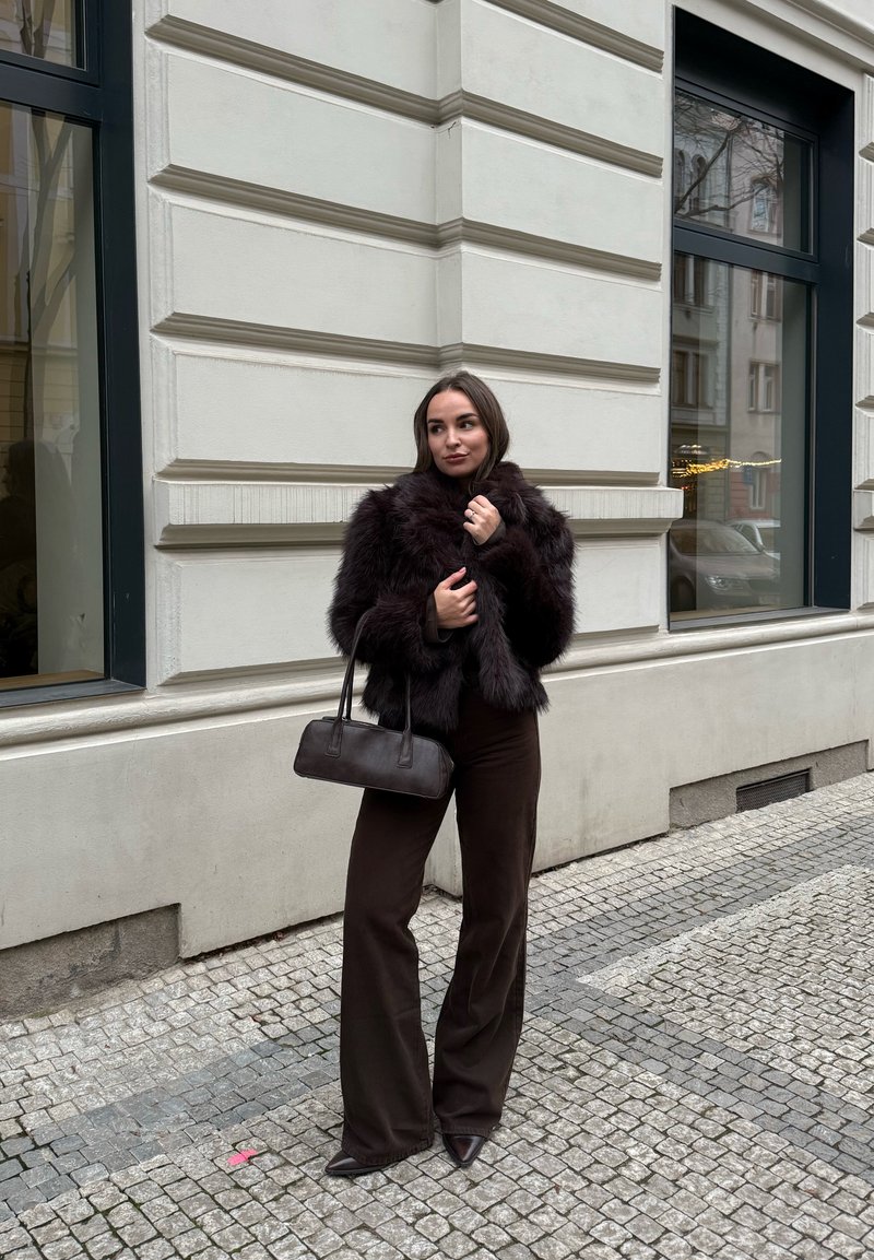 Femme debout sur une rue pavée portant une veste en fourrure foncée, un pantalon à jambes larges, des chaussures pointues, et tenant un petit sac à main en cuir près d'une façade de bâtiment.
