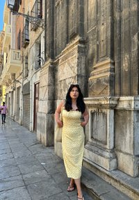 Κίτρινο φόρεμα με κόκκινες λουλουδάτες λεπτομέρειες, εφαρμοστή σχεδίαση, μέσου μήκους. Υφασμάτινη τσάντα και μαύρες παντόφλες. Τοίχος από πέτρα στο φόντο με αρχιτεκτονικές λεπτομέρειες.