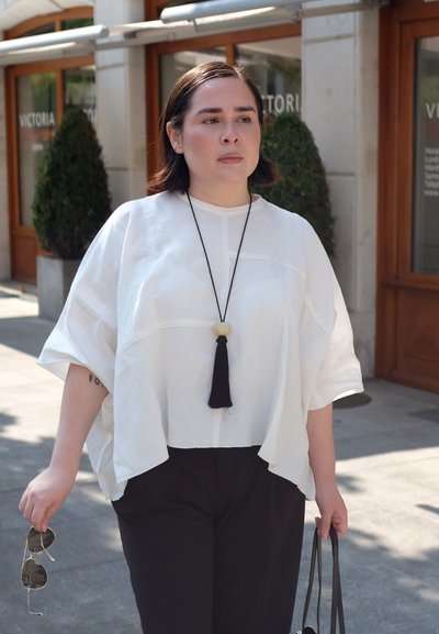 Mujer caminando al aire libre con una blusa blanca holgada y pantalones negros, sosteniendo gafas de sol y un bolso negro, cerca de la entrada de un edificio.