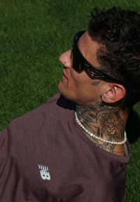 Camiseta corta de color marrón con un logo blanco, gafas de sol negras, múltiples cadenas y un collar de perlas adornan a una persona con intrincados tatuajes.