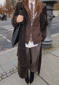 Barna szarvasbőrkabát barna cipzáras pulóver fölött, fehér inggel és krémszínű kockás nyakkendővel. Szürke bő szárú nadrág és fekete hegyes orrú csizma. Fekete bevásárlótáska.