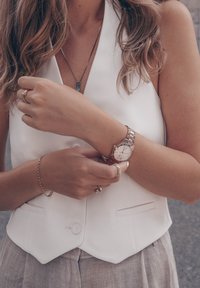 Silver watch with a white face and rose gold accents. Worn on a wrist alongside rings and a thin gold bracelet. Model in a white vest.