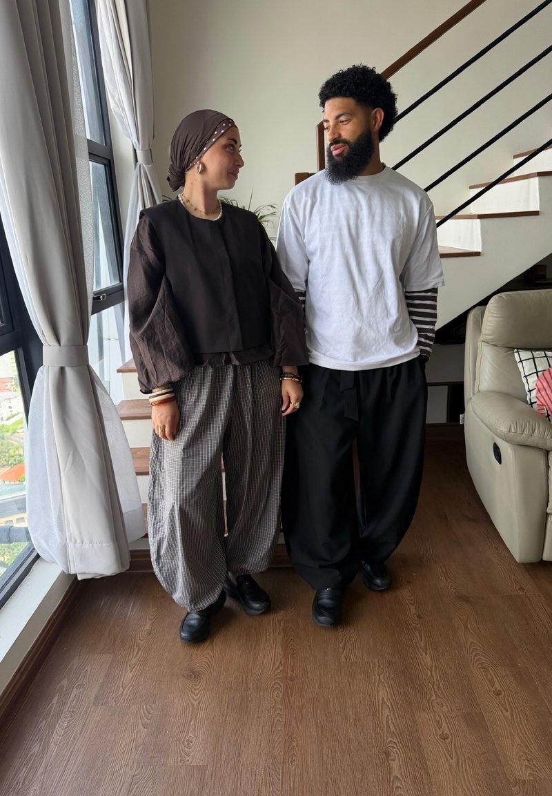 Une femme portant un foulard et des vêtements amples se tient à côté d’un homme barbu portant plusieurs couches de hauts, tous deux se regardant à l’intérieur près d’un escalier.