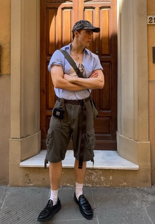 Joven con gorra, camiseta de manga corta, pantalones cortos cargo, calcetines blancos y zapatos negros, de pie con los brazos cruzados frente a una puerta de madera.