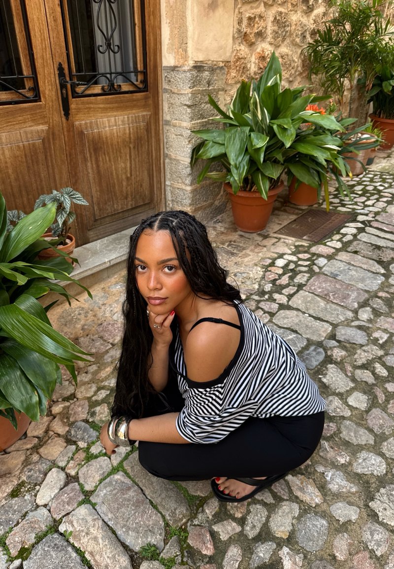 Femme aux longs cheveux tressés accroupie sur une rue pavée près d'une porte en bois et de plantes vertes en pot, portant un haut rayé et un pantalon noir.