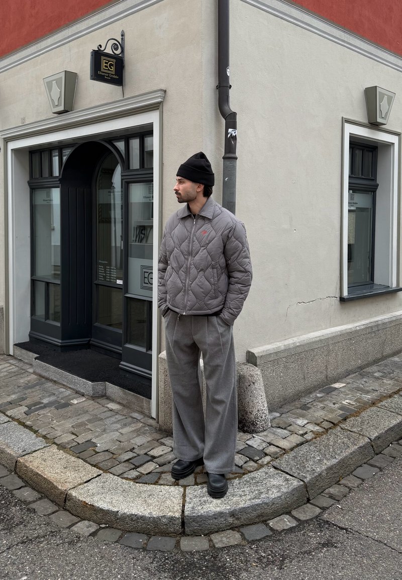 Muž v sivej prešívanej bunde, čiernej čiapke a sivých nohaviciach stojí na rohu chodníka z kociek pred vchodom do béžovej budovy.