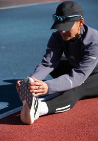 Kvinde i sportstøj der strækker sit ben på en udendørs løbebane, mens hun sidder på den røde og blå baneoverflade og holder om sin fod.