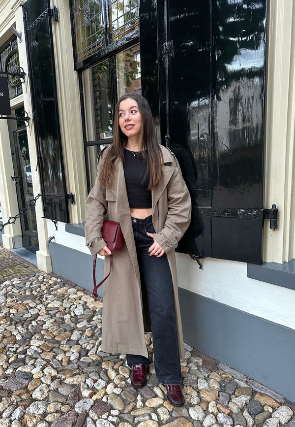 Helles beiges Trenchcoat über einem schwarzen Crop-Top, kombiniert mit schwarzen Jeans und burgundy Schuhen. Hält eine kleine burgundy Tasche. Kopfsteinpflasterboden.