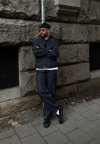 Άνδρας με μαύρο σκουφί, γυαλιά ηλίου και σκούρα τζην μπουφάν και παντελόνι στέκεται με τα χέρια σταυρωμένα, ακ leaning against a textured stone wall.