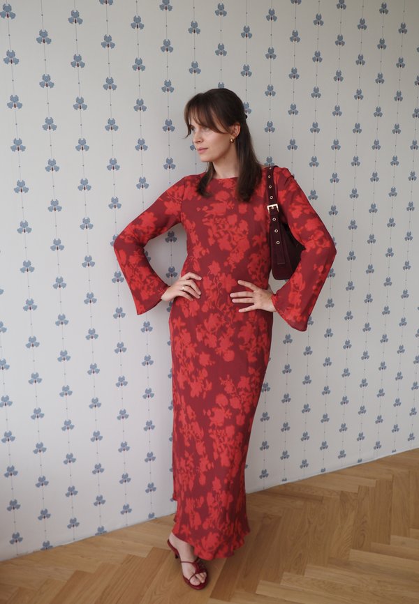 Mujer con un vestido largo rojo con flores y sandalias posando con las manos en las caderas frente a una pared blanca con un patrón azul.