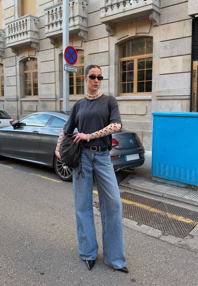 Camiseta negra con mangas largas recortadas, jeans de pierna ancha azul claro, tacones de cuña negros y un bolso negro. Los accesorios incluyen un collar de perlas y gafas de sol.