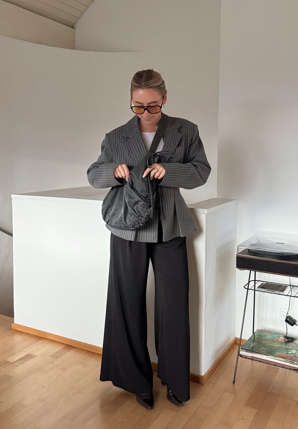 Mujer con blazer de rayas de gran tamaño y pantalones negros anchos, ajustando bolso negro de hombro, de pie en el interior cerca de paredes blancas y suelo de madera.