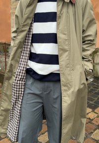 Beige outerwear coat with a check interior, worn over a navy and white striped jumper, paired with grey trousers.