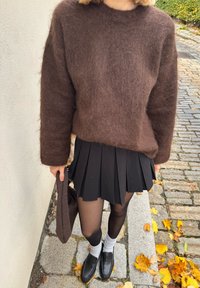 Suéter castanho felpudo, saia plissada preta, collants pretos, meias brancas e mocassins pretos. Asegurando uma mala castanha escura. Fundo de calçada de pedras.