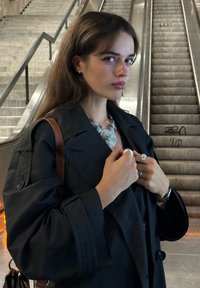 Palton negru cu rever larg, ținându-se închis cu material. Colier circular din argint și mai multe inele. Textură netedă, design minimalist.