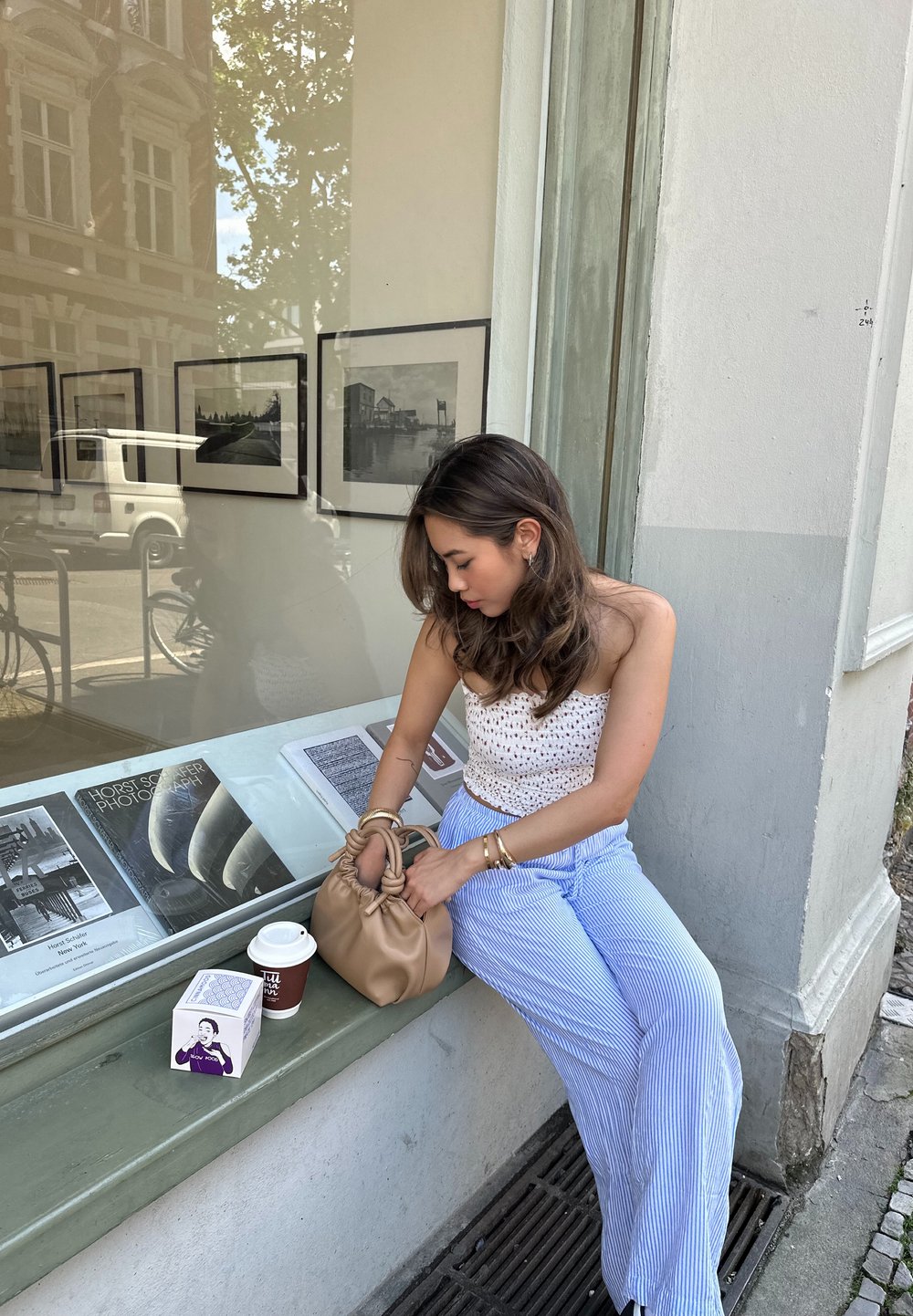 Μπεζ τσάντα με λεπτομέρεια κόμπου, λευκό μπλουζάκι με μικροσκοπικό λουλούδι, μπλε και λευκές ρίγες παντελόνι σε φαρδιά γραμμή, φλιτζάνι καφέ και ένα κουτί σε μια πέτρινη προεξοχή.