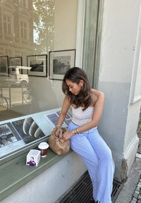 Μπεζ τσάντα με λεπτομέρεια κόμπου, λευκό μπλουζάκι με μικροσκοπικό λουλούδι, μπλε και λευκές ρίγες παντελόνι σε φαρδιά γραμμή, φλιτζάνι καφέ και ένα κουτί σε μια πέτρινη προεξοχή.