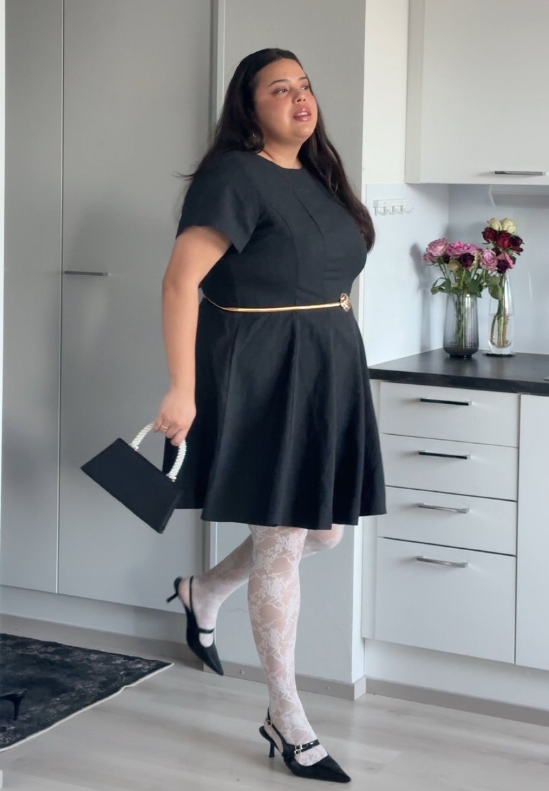 Femme en robe noire avec ceinture dorée, collants blancs à motifs floraux et talons noirs tenant un petit sac à main noir dans une cuisine moderne.