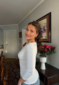 Jeune femme aux cheveux foncés attachés en queue de cheval, souriante, vêtue d'un haut blanc et d'un jean, debout dans une pièce avec un pot de fleurs et une œuvre d'art colorée encadrée.