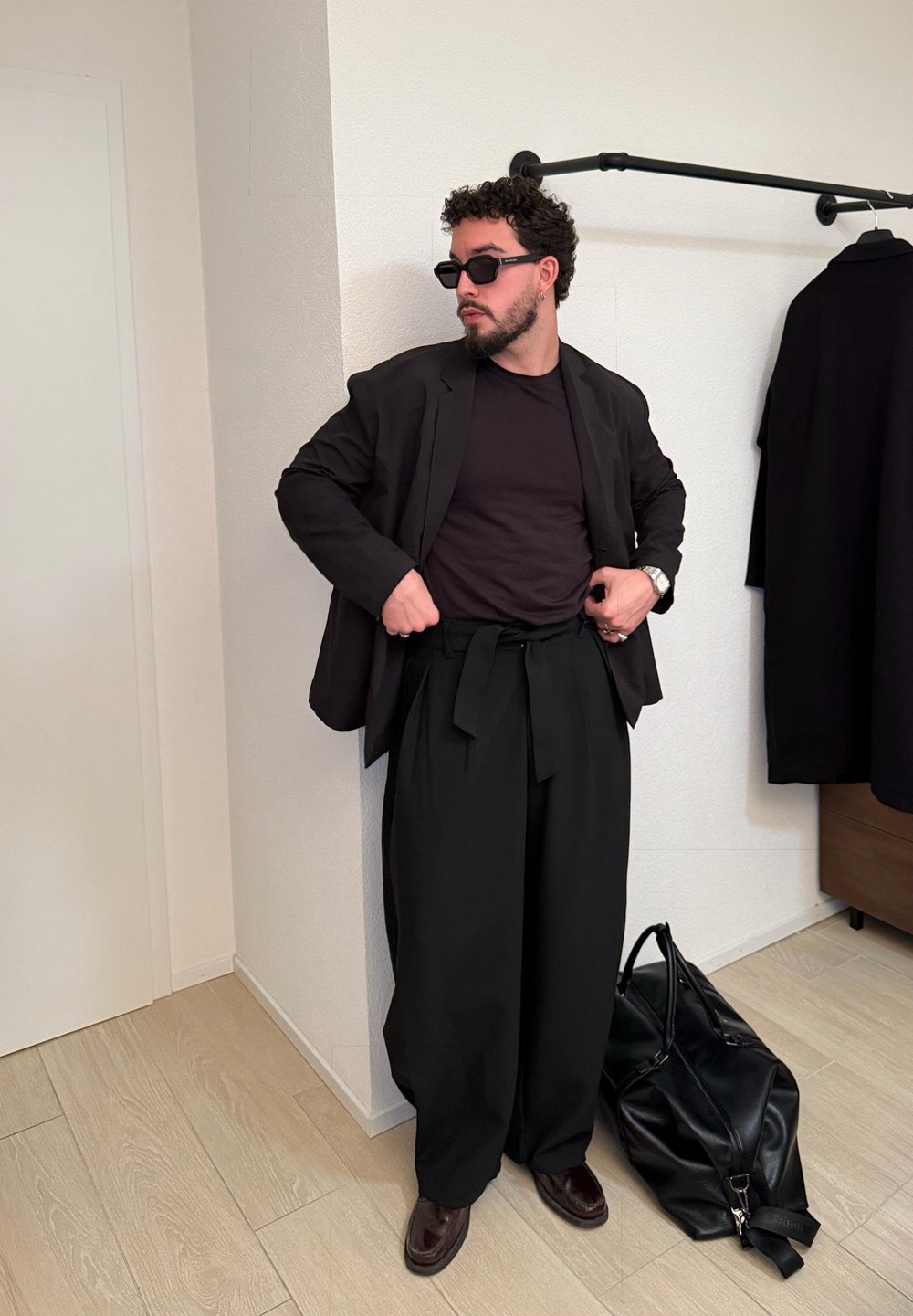 Homme portant un blazer noir, un pantalon noir ample, des lunettes de soleil et des chaussures en cuir, ajustant son pantalon dans une pièce avec un manteau noir suspendu et un grand sac noir.