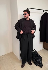 Homme portant un blazer noir, un pantalon noir ample, des lunettes de soleil et des chaussures en cuir, ajustant son pantalon dans une pièce avec un manteau noir suspendu et un grand sac noir.