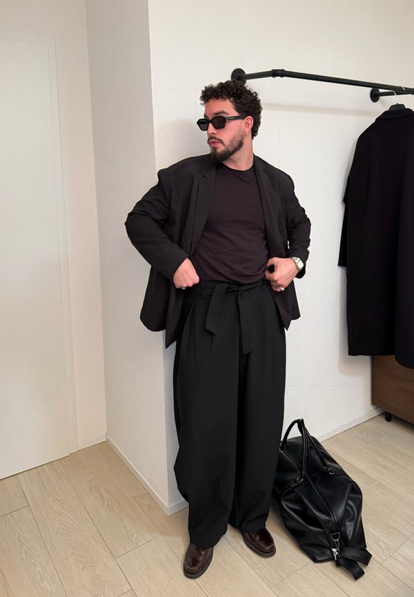 Uomo che indossa un blazer nero, pantaloni neri larghi, occhiali da sole e scarpe di pelle mentre sistema i pantaloni in una stanza con un cappotto nero appeso e una grande borsa nera.