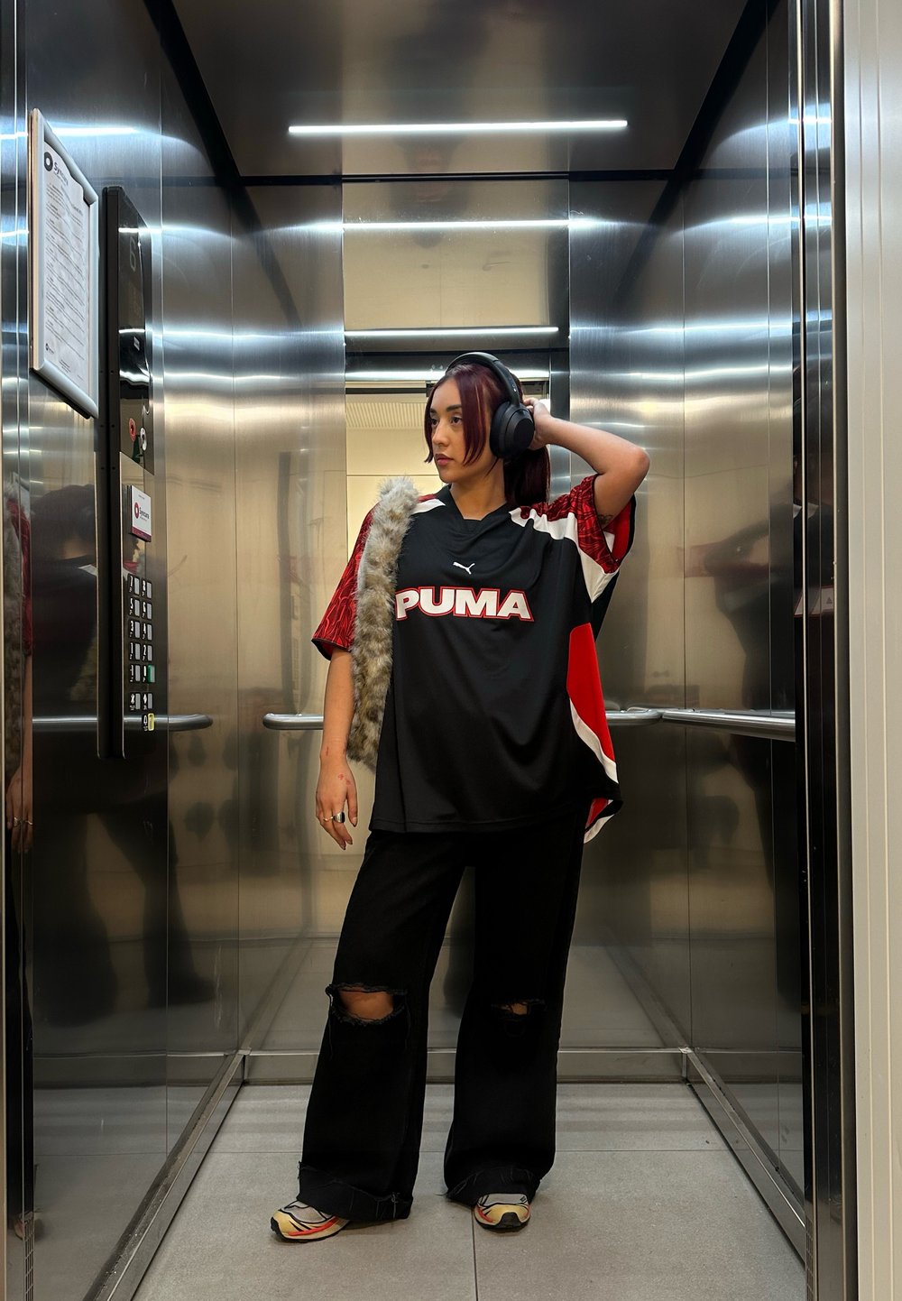 PUMA oversized černý dres s červenými akcenty, spárovaný s potrhanými černými džíny a vzorovanými teniskami, v metalovém výtahu.