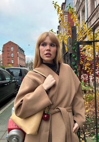 Manteau en laine beige avec une ceinture, finition texturée, associé à un sac à main en cuir jaune orné d'un porte-clés en forme de pomme rouge. Fond urbain.