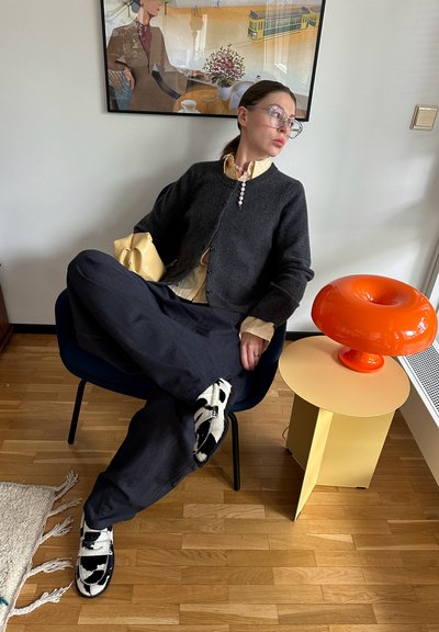 Cardigan negro de lana sobre una camiseta de rayas amarillas, pantalones azules y zapatos con estampado de vaca. Lámpara decorativa naranja sobre una mesa lateral amarilla, suelo de madera.