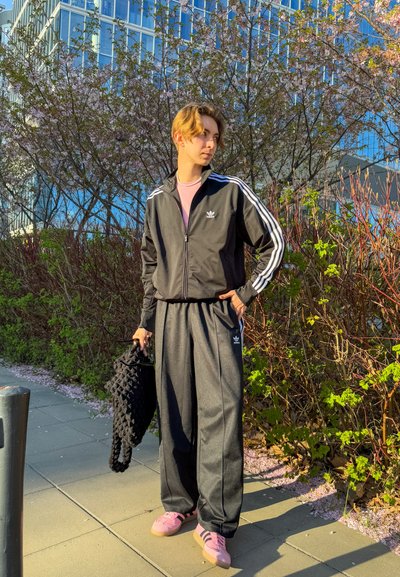 Joven vestido con un chándal negro de Adidas y zapatillas rosas, de pie en una acera urbana, sosteniendo una bolsa de mimbre negra cerca de arbustos en flor.