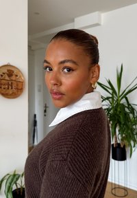 Jonge vrouw met een strakke bun, gouden ringoorbellen en een bruine trui over een witte blouse, staande binnenshuis bij planten en wanddecoraties.