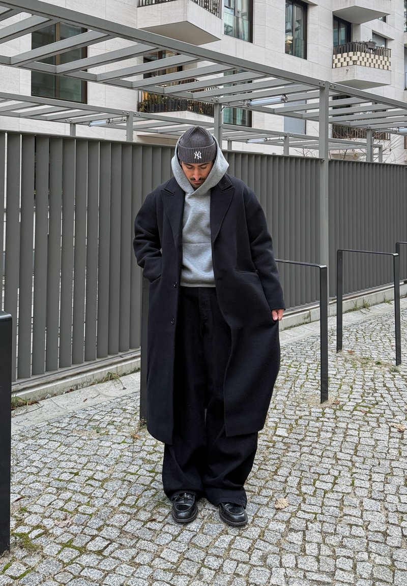 Homme portant un pardessus noir, un sweat à capuche gris, un bonnet noir et un pantalon noir ample, debout sur un chemin pavé près d'un bâtiment moderne.