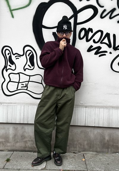 Chaqueta de forro polar burdeos con cuello alto, pantalones de corte ancho en verde oliva, gorro negro con logo blanco y zapatos oscuros sobre una superficie texturizada.