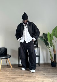Jachetă bomber neagră peste o cămașă albă, asortată cu pantaloni largi negri și adidași albi. Accent de o căciulă neagră cu textură. Mediu interior.