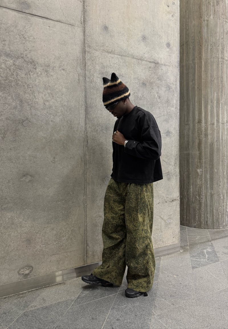 Schwarzes, oversized Langarmshirt, grüne, gemusterte weite Hose und eine gestreifte Mütze mit Ohren. Schwarze Schuhe. Texturierter Beton-Hintergrund.