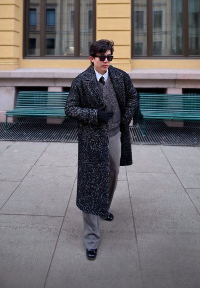 Hombre con abrigo largo oscuro con estampado, pantalones grises, guantes, gafas de sol y corbata camina junto a bancos verdes en un entorno urbano.