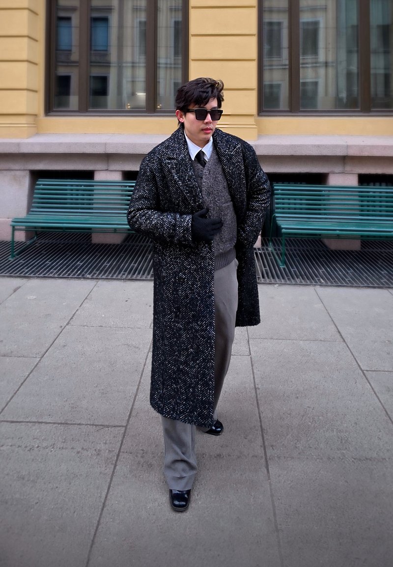 Homme portant un long manteau foncé à motifs, un pantalon gris, des gants, des lunettes de soleil et une cravate, marchant devant des bancs verts dans un cadre urbain.