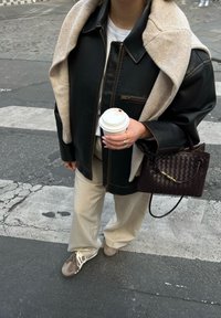 Giacca di pelle nera con un maglione beige drappeggiato sulle spalle, t-shirt bianca, pantaloni beige a gamba larga, borsa intrecciata marrone e sneakers taupe.