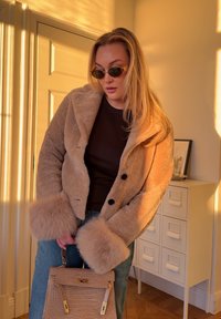 Krótkie beżowe futerko ze sztucznego futra z czarnymi guzikami i futrzanymi mankietami. Modelka trzyma teksturowaną, jasnoróżową torebkę, nosząc ciemne okulary przeciwsłoneczne.