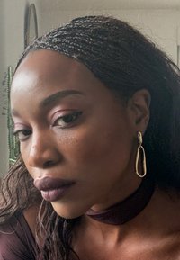 Pendiente de aro ovalado dorado, piel morena, cabello rizado oscuro, labios burdeos intensos, sombra de ojos rosa suave, camiseta oscura con escote tipo gargantilla.