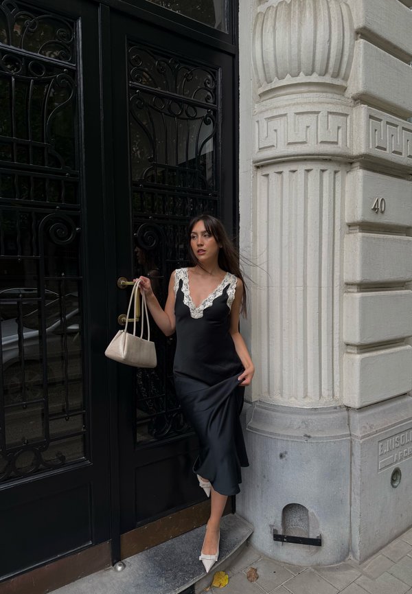 Femme en robe en satin noir avec bordure en dentelle tenant un sac beige, debout près d'une porte noire ornée et d'une colonne blanche portant le numéro 40.