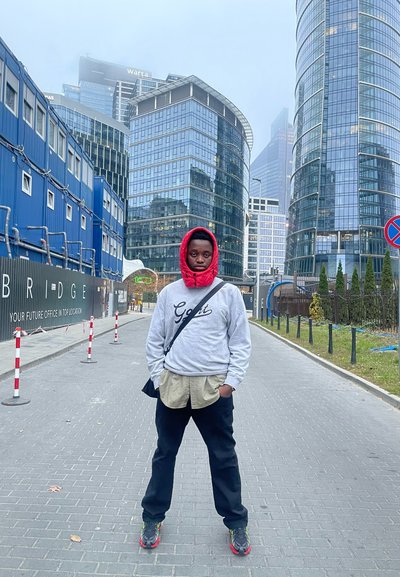 Individuo que lleva una chaqueta con capucha roja sobre un sudadera gris y una camisa de botones beige, pantalones negros y zapatillas coloridas, de pie en una zona urbana.