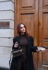Zwarte wollen overjas, bruin aansluitend top, bruine broek. Houdt een kleine bruine tas met een rits vast en een afhaal koffie in een kartonnen houder.