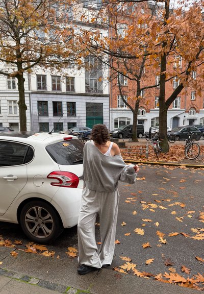 Sudadera gris claro extragrande y pantalones deportivos a juego, hechos de tela suave. Zapatos negros. Fondo con hojas otoñales y edificios.