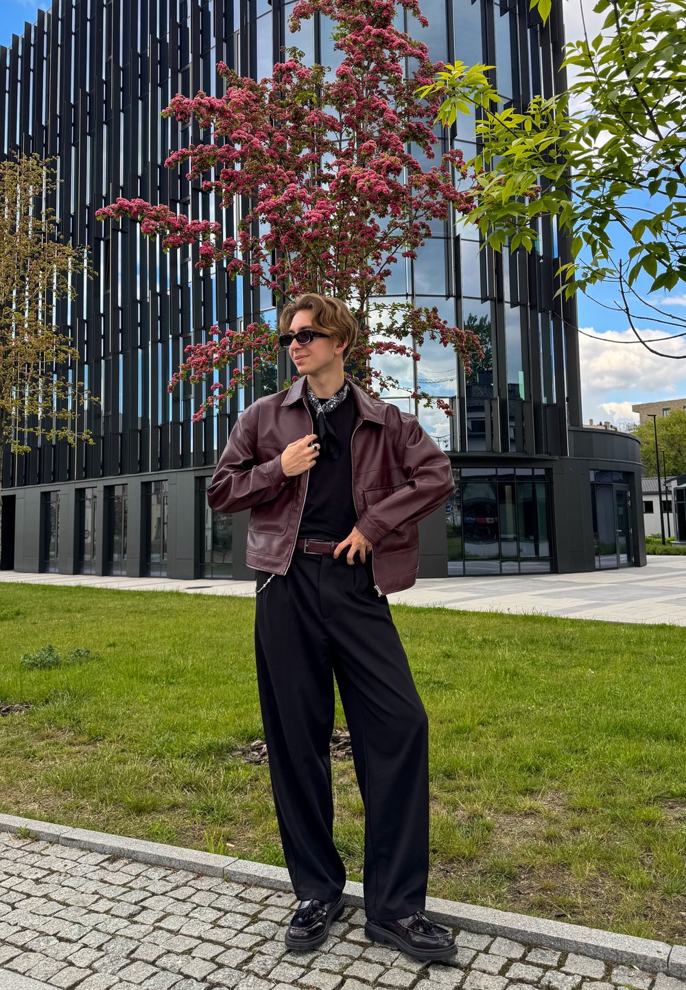 Jaqueta de couro castanha, camisa preta, calças largas pretas, sapatos pretos. Cenário ao ar livre com vegetação e um edifício moderno ao fundo.