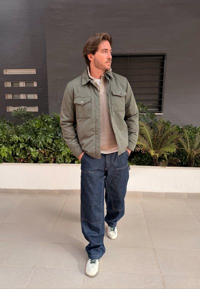 Hombre caminando en interiores con chaqueta verde, suéter beige, pantalones azul oscuro holgados y zapatillas blancas con las manos en los bolsillos.