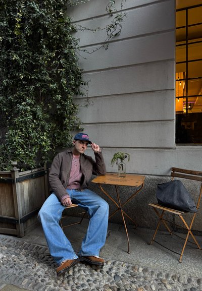 La ropa informal incluye una chaqueta marrón, una camiseta a rayas rojas y blancas, y jeans de color azul claro. Una mesa sostiene un jarrón de cristal con una planta.
