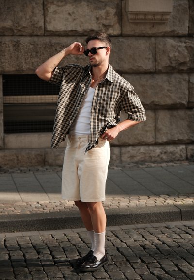 Conjunto informal que incluye una camisa de cuadros negra y beige de manga corta, una camiseta blanca sin mangas, pantalones cortos beige y zapatos negros con calcetines blancos.