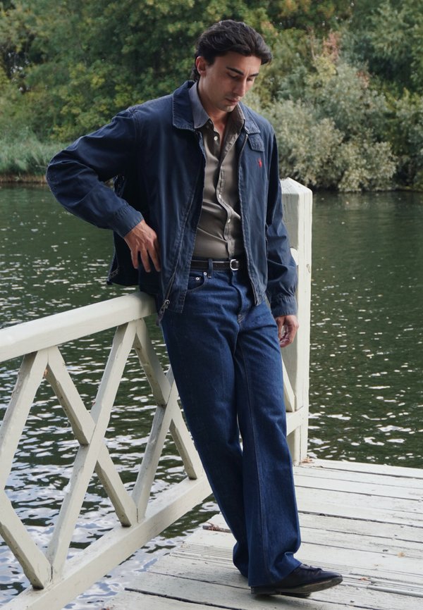 Uomo con giacca blu e jeans che si appoggia a una ringhiera bianca di legno vicino a un lago con alberi sullo sfondo, guardando in basso con aria pensierosa.
